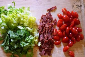 Chopped lettuce, bacon and grape tomatoes sitting in rows on a wooden cutting board.
