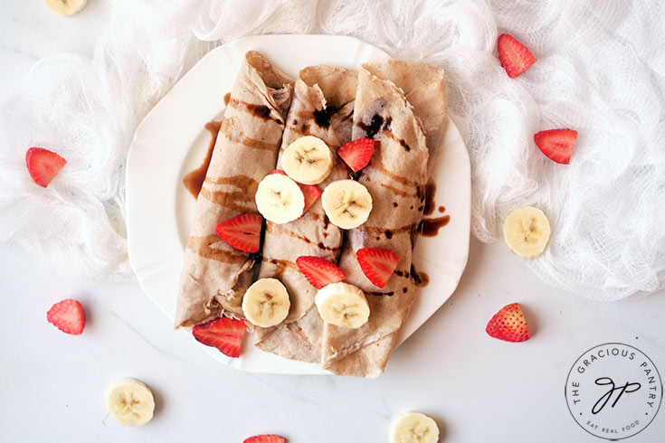 An overhead shot looking down onto a plate of rolled crepes, topped with sliced strawberries and bananas.