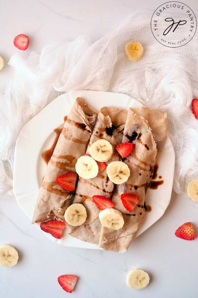 An overhead view looking down on a white, round plate holds three rolled crepes that are topped with sliced banana, strawberries and drizzled with some chocolate syrup.