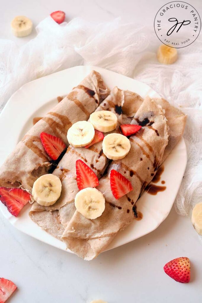 A white, round plate holds three rolled crepes that are topped with sliced banana, strawberries and drizzled with some chocolate syrup.