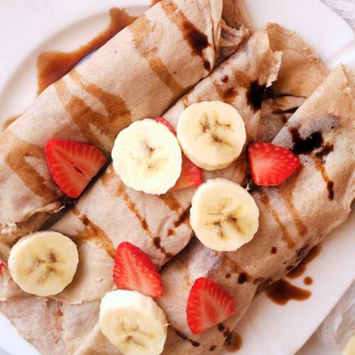 A close up, overhead shot of rolled crepes topped with sliced strawberries, bananas an a chocolate drizzle.