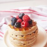 A side view of a stack of buckwheat pancakes sitting on a white plate, topped with fresh berries and maple syrup.