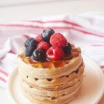 A side view of a stack of buckwheat pancakes sitting on a white plate, topped with fresh berries and maple syrup.