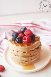 A side view of a stack of buckwheat pancakes sitting on a white plate, topped with fresh berries and maple syrup.