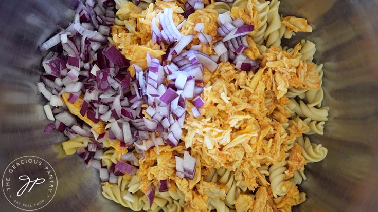 Chopped red onions, shredded buffalo chicken and cooked rotini pasta in a mixing bowl.