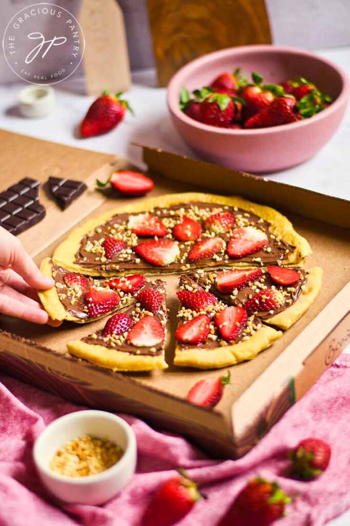 A hand reaches for a slice of chocolate pizza sitting on a wooden cutting board.