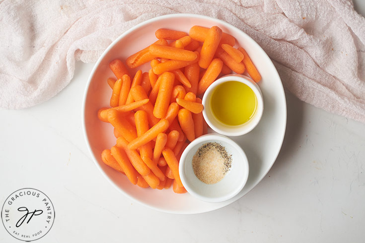 Baby carrots in a white bowl with two white ramekins holding oil and spices.
