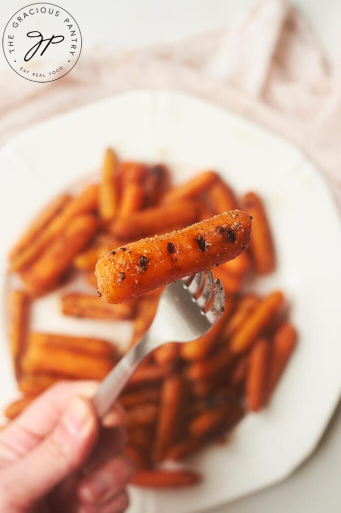 A fork holds a single grilled baby carrot towards the camera, with a white plate filled with grilled baby carrots sitting on the table behind it.
