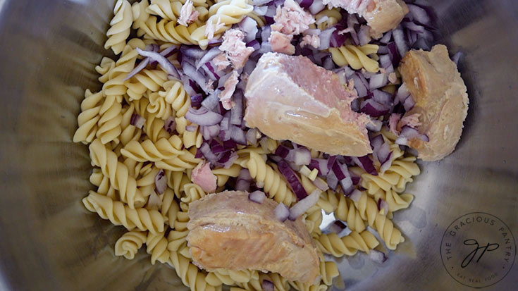 Canned tuna added to pasta and onions in a mixing bowl.