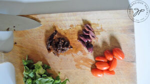 Lettuc, kalamata olives, sun dried tomatoes and fresh grape tomatoes sitting in small piles on a cutting board.