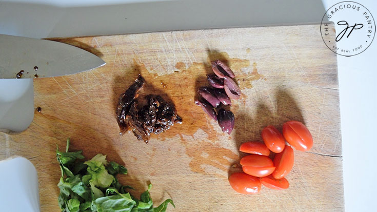 Lettuc, kalamata olives, sun dried tomatoes and fresh grape tomatoes sitting in small piles on a cutting board.