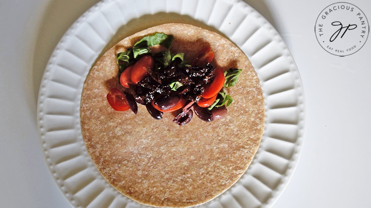 Lettuce. tomatoes, olives and sun dried tomatoes piled onto one side of a whole grain tortilla which is laying on a white plate.