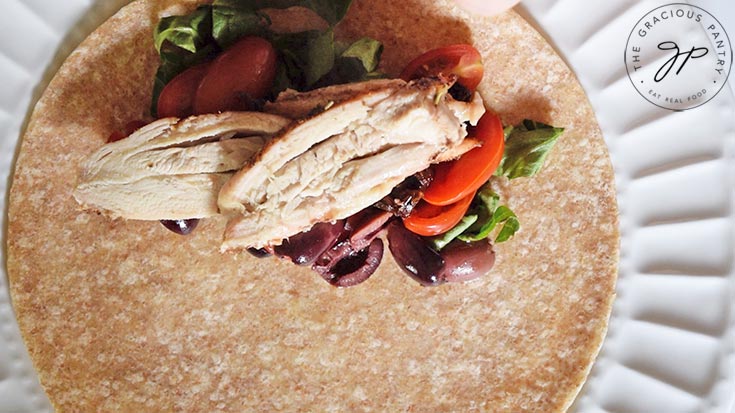 Chicken slices layered over vegetables on a whole grain tortilla which will get rolled into an Italian chicken wrap.