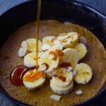 Maple syrup being poured over a skillet pancake topped with sliced bananas and sliced almonds.