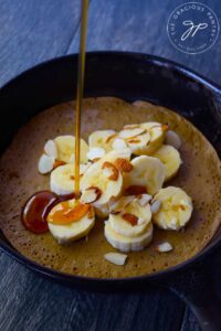 Maple syrup being poured over a skillet pancake topped with sliced bananas and sliced almonds.