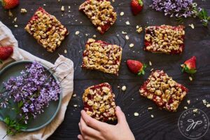 A hand reaches for a healthy strawberry crumb bar on a dark gray surface.