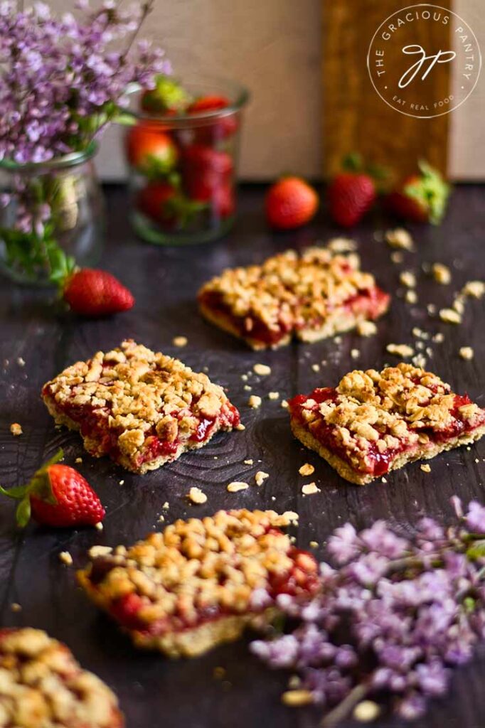 A group of four healthy strawberry crumb bars sit on a gray surface with a vase of purple flowers and a jar of fresh strawberries sitting behind them.