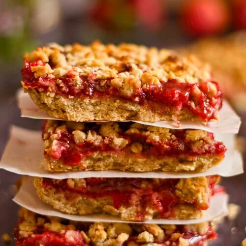 A stack of healthy strawberry crumb bars sit on a dark surface with pieces of parchment between each bar.