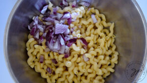 Adding chopped red onion to cooked and cooled pasta in a large stainless steel mixing bowl.