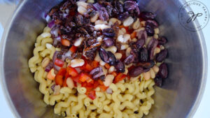 Sun-dried tomatoes and olives added to peppers, onions and cooked pasta in a mixing bowl.