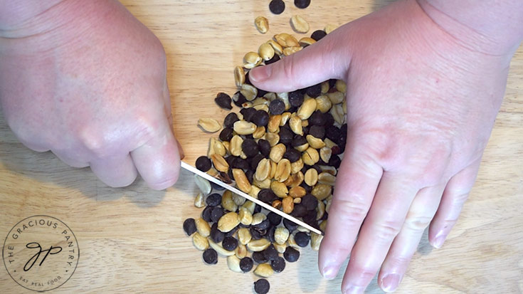Hands chopping the peanuts and chocolate chips on a wooden cutting board.