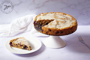 A Blueberry Cake on a cake stand with one piece removed and sitting on a white plate next to it.