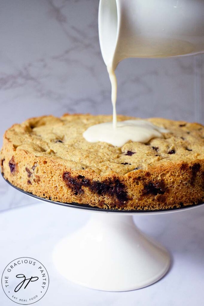 Lemon icing being poured from a white pitcher, over a baked blueberry cake.