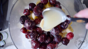 Pouring lemon juice onto frozen fruit sitting in a food processor.