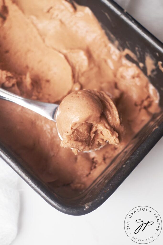 An ice cream scoop lays inside a bread pan filled with chocolate ice cream. It holds a scoop of that ice cream.