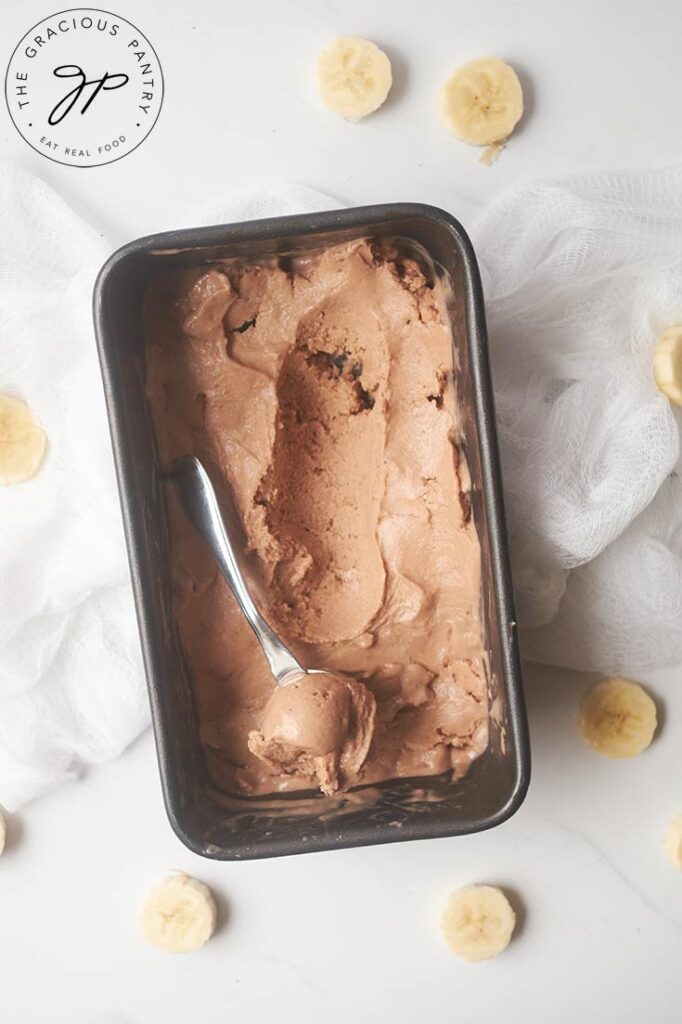 An ice cream scooper lays in a loaf pan filled with Chocolate Banana Nice Cream, and holds a scoop of the ice cream.