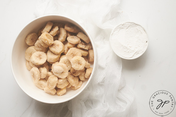 Two white bowls on a white surface. One filled with frozen banana slices, the other with partially frozen coconut milk.