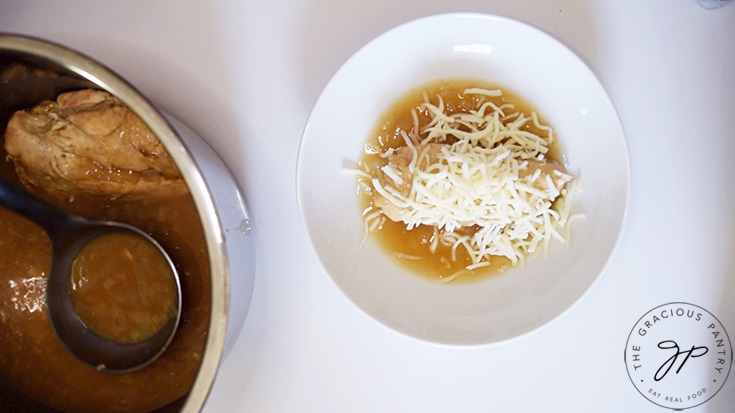 A chicken breast with french onion soup in a bowl and topped with grated cheese.