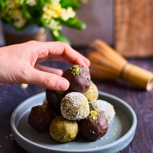 A female hand reaches for a matcha energy ball from a plate stacked high with them.