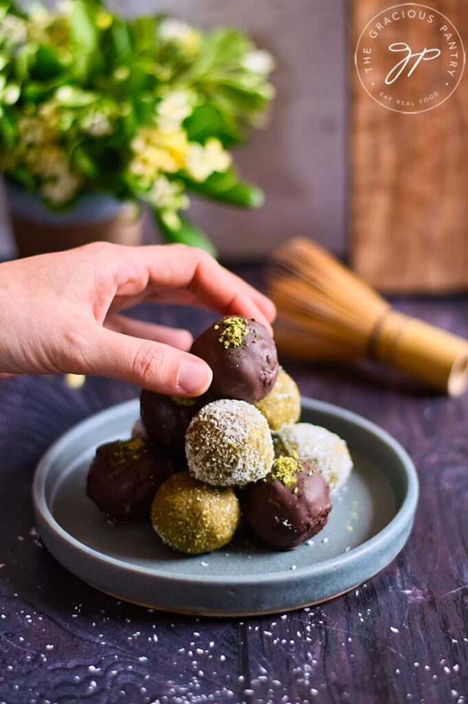 A female hand reaches for a matcha energy ball from a plate stacked high with them.