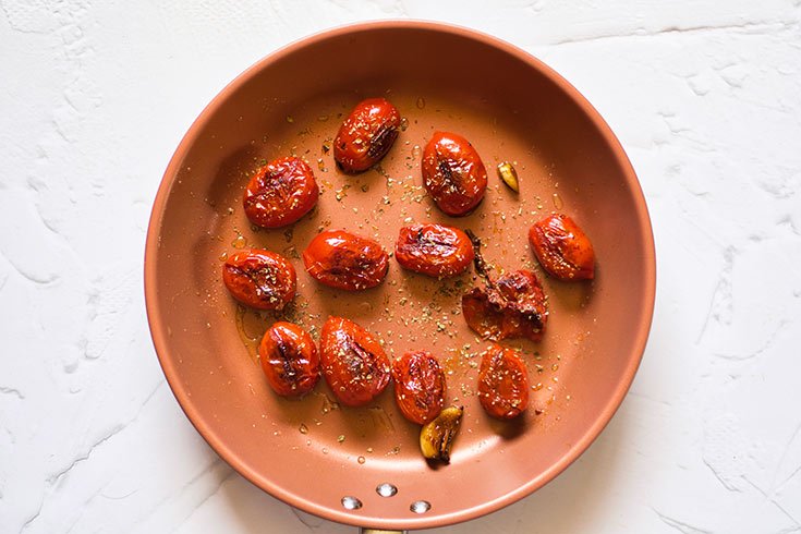 Sautéd, whole grape tomatoes and a garlic clove sitting slightly charred in a skillet.