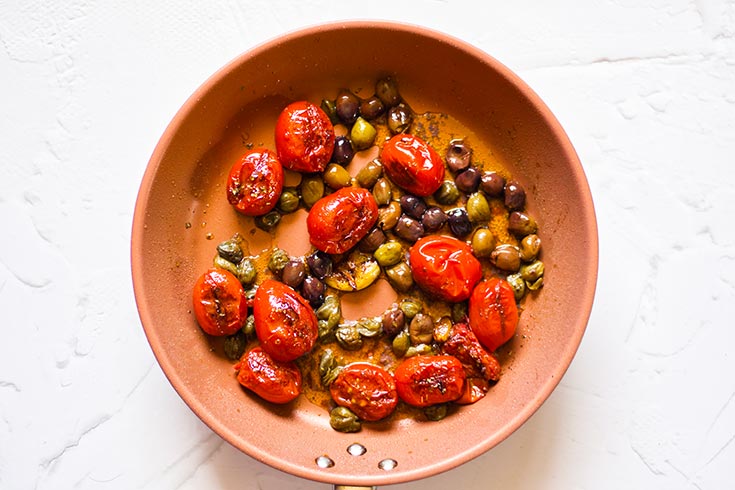 Charred grape tomatoes, a garlic clove and capers sitting in olive oil in a skillet.