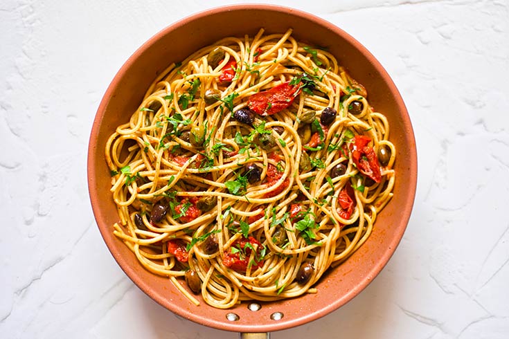 Pasta stirred into the tomatoes and capers with chopped olives added and sitting in a skillet.