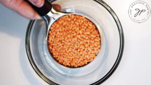 A hand holds a measuring cup filled with red lentils over a glass mixing bowl.