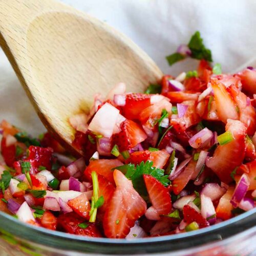 A glass bowl holds fresh strawberry salsa. A wooden spoon sits against the inside of the bowl.