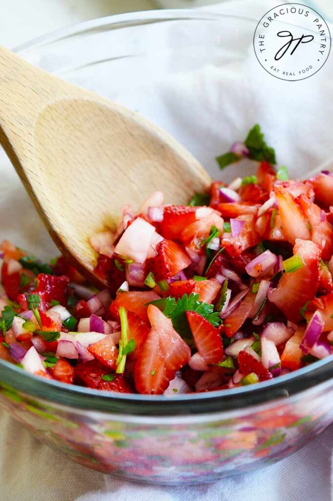 A glass bowl holds fresh strawberry salsa. A wooden spoon sits against the inside of the bowl.