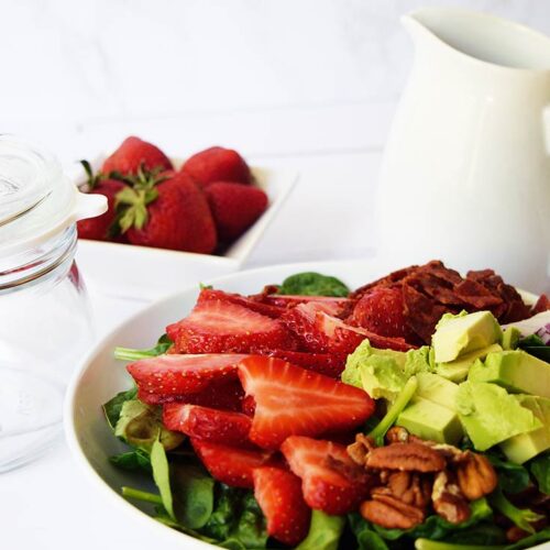 A white bowl sits on a white surface and is filled with Strawberry Spinach Salad.