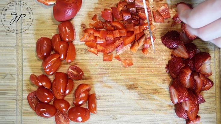 A cutting board with sliced tomatoes, strawberries and chopped red peppers.