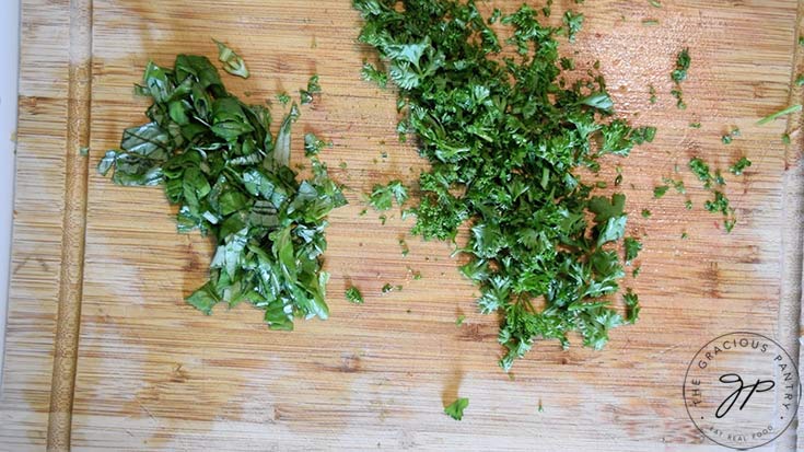 A wooden cutting board with chopped fresh basil and parsley.