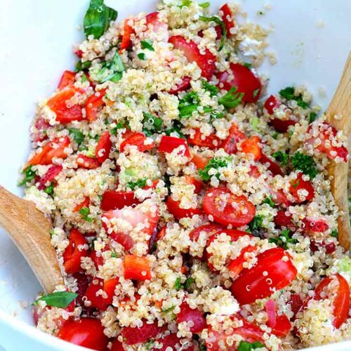 A close look at Summer Quinoa Salad sitting in a white mixing bowl with two wooden spoons.