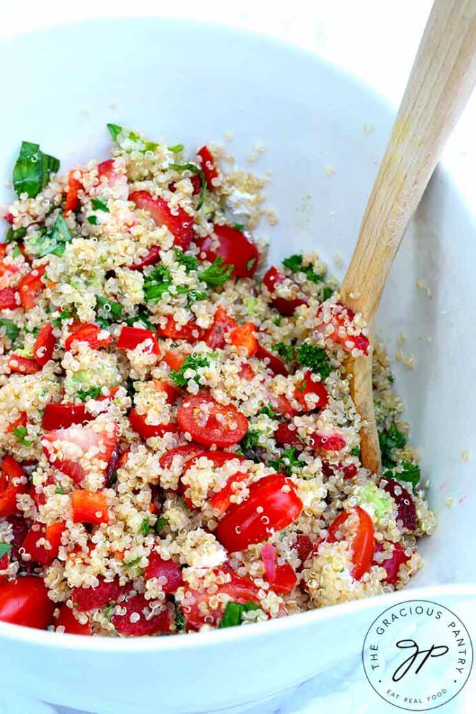 A close up view of this Summer Quinoa Salad Recipe sitting in a white mixing bowl with a wooden spoon.