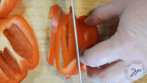 A red bell pepper being sliced on a wooden cutting board.