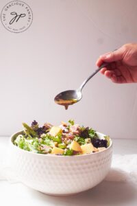 A spoon of dressing being drizzled over a white bowl filled with Chicken Apple Salad.