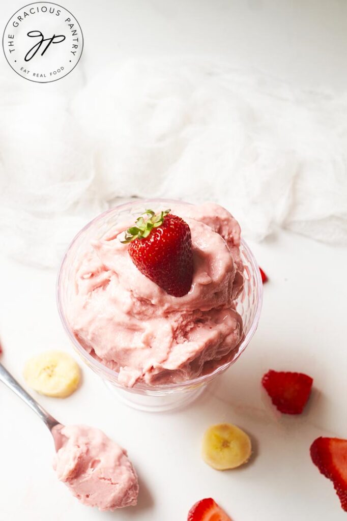 An overhead side view of a glass dish holding some Healthy Strawberry Ice Cream. A spoon of ice cream rests to the left side of the dish.