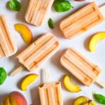 Homemade Peach Popsicles lay arranged on a white surface with fresh peaches.