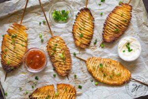 Cooked accordion potatoes on skewers laying on parchment with small bowls of condiments around them.